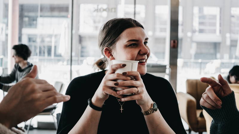 Woman holding cup at a coffee house talking to friends about how much they make